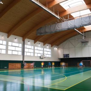 Campo sportivo indoor multifunzionale con tetto in legno e alcuni giocatori sul campo, visti da lontano