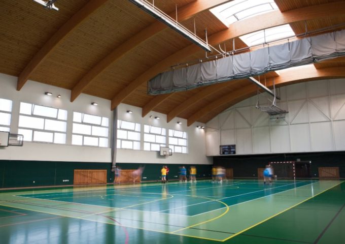 Campo sportivo indoor multifunzionale con tetto in legno e alcuni giocatori sul campo, visti da lontano