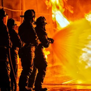 Quattro Vigili del Fuoco altamente equipaggiati sostengono e usano una manichetta provando a domare con l'acqua un grande incendio