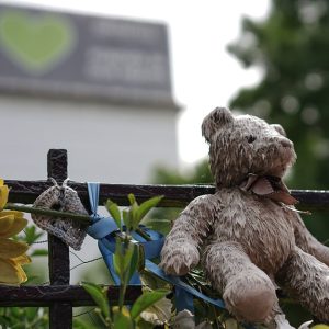 Un cancello nei pressi della Grenfell Tower, che si vede sullo sfondo, con fiori e oggetti in ricordo delle vittime