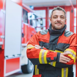 Un veterano dei Vigili del Fuoco a braccia conserte e sorridente all'interno di una caserma, accanto a un'autopompa