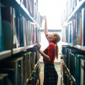 Una giovane bibliotecaria con golfino rosso e gonna a scacchi sta prendendo un libro sui ripiani alti di uno scaffale nello stretto corridoio di una biblioteca