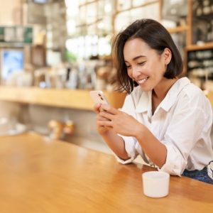 Una ragazza orientale sorride mentre guarda il suo smartphone seduta su un lungo tavolo in una caffetteria in stile anglosassone con arredi in legno