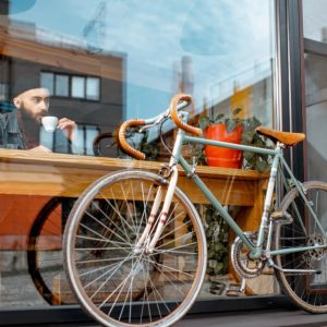 Vista dall'esterno di una vetrina di un locale, con una bicicletta da corsa appoggiata sulla vetrina all'esterno e, dentro, un ragazzo con la barba lunga e lo stile hipster che sta seduto a un tavolino