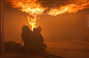 Due vigili del fuoco equipaggiati con respiratore e bombole stanno intervenendo accucciati a terra in una stanza piena di fiamme e fumo