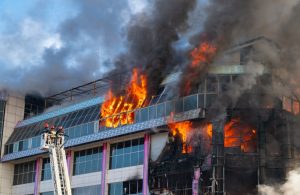 Un grande edificio commerciale o industriale con le fiamme che escono da alcune grandi finestre vetrate, mentre i vigili del fuoco stanno intervenendo su di un'autoscala