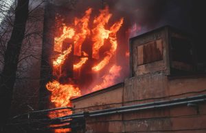 Primo piano su un vecchio edificio industriale con, in secondo piano, un alto palazzo che brucia, con le fiamme che escono dalle finestre
