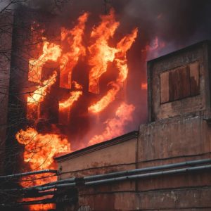 Primo piano su un vecchio edificio industriale con, in secondo piano, un alto palazzo che brucia, con le fiamme che escono dalle finestre