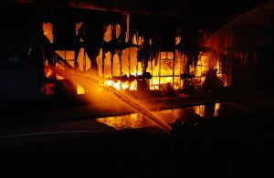 Lo scheletro di un edificio quasi totalmente incendiato nella notte, con le fiamme che avvolgono tutto e il getto d'acqua dei pompieri diretto sulle fiamme