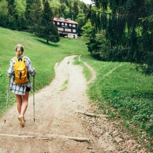 Una ragazza bionda in pantaloncini, vista di spalle, sta camminando con i bastoni da trekking lungo un sentiero che porta a un rifugio alpino tra i boschi