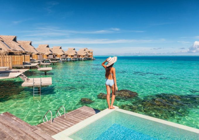 Una donna in costume con un grande cappello, vista di spalle in piedi sul bordo di una piscina di un villaggio turistico, con l'acqua cristallina del mare a pochi passi, e una fila di bungalow su palafitte