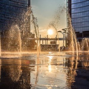 Vista di Porta Garibaldi, a Milano, di tardo pomeriggio, con il sole calante che crea effetti visivi sugli spruzzi d'acqua di una fontana