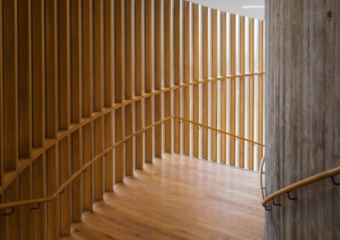 Uno spazio architettonico interno con una scala curva in legno dotata di corrimano su entrambi i lati. La scala è racchiusa da doghe verticali di legno su un lato e da un muro di cemento sull'altro. Le doghe in legno permettono alla luce di filtrare attraverso, creando un interessante gioco di luci e ombre sul pavimento e sulle pareti. Il pavimento e i corrimano sono realizzati nello stesso legno