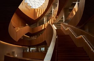 L'interno di una biblioteca pubblica moderna caratterizzata da una grande scala a chiocciola in legno. La scala presenta linee eleganti e curve ed è illuminata dalla luce naturale proveniente da un lucernario geometrico sopra di essa. Il design include diversi livelli con ringhiere in legno e pannelli di vetro, creando un'atmosfera aperta e ariosa