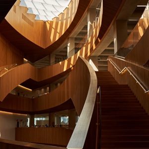 L'interno di una biblioteca pubblica moderna caratterizzata da una grande scala a chiocciola in legno. La scala presenta linee eleganti e curve ed è illuminata dalla luce naturale proveniente da un lucernario geometrico sopra di essa. Il design include diversi livelli con ringhiere in legno e pannelli di vetro, creando un'atmosfera aperta e ariosa