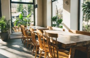 L'interno di un ristorante moderno e luminoso. La stanza è caratterizzata da grandi finestre che permettono alla luce naturale di inondare lo spazio, creando un'atmosfera accogliente. Al centro, c'è un lungo tavolo di legno con diverse sedie di legno disposte intorno. Numerose piante in vaso sono posizionate intorno al locale, aggiungendo un tocco di verde e freschezza. Le finestre offrono una vista su un'area esterna con altre piante e alberi visibili