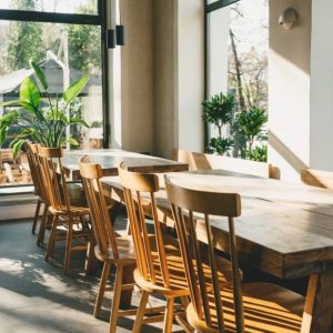 L'interno di un ristorante moderno e luminoso. La stanza è caratterizzata da grandi finestre che permettono alla luce naturale di inondare lo spazio, creando un'atmosfera accogliente. Al centro, c'è un lungo tavolo di legno con diverse sedie di legno disposte intorno. Numerose piante in vaso sono posizionate intorno al locale, aggiungendo un tocco di verde e freschezza. Le finestre offrono una vista su un'area esterna con altre piante e alberi visibili