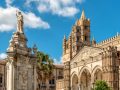 La Cattedrale di Palermo. In primo piano, c'è una statua monumentale di una figura religiosa che tiene una croce, posta su un piedistallo decorato con dettagli architettonici elaborati. Sullo sfondo, si può vedere la facciata della cattedrale, caratterizzata da archi gotici, intricate sculture e un campanile imponente. La cattedrale è un esempio significativo di architettura normanna e gotica