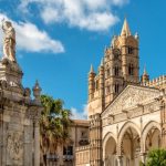 La Cattedrale di Palermo. In primo piano, c'è una statua monumentale di una figura religiosa che tiene una croce, posta su un piedistallo decorato con dettagli architettonici elaborati. Sullo sfondo, si può vedere la facciata della cattedrale, caratterizzata da archi gotici, intricate sculture e un campanile imponente. La cattedrale è un esempio significativo di architettura normanna e gotica