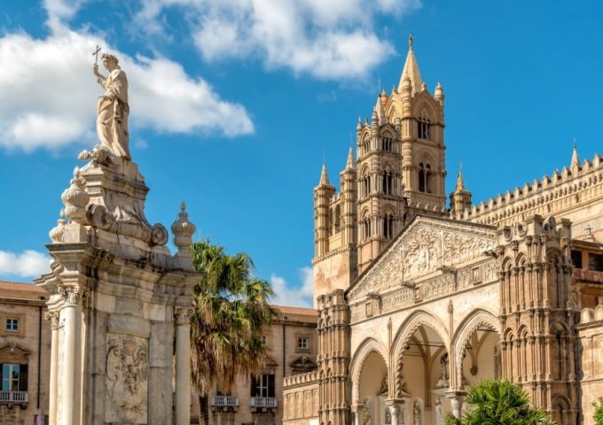 La Cattedrale di Palermo. In primo piano, c'è una statua monumentale di una figura religiosa che tiene una croce, posta su un piedistallo decorato con dettagli architettonici elaborati. Sullo sfondo, si può vedere la facciata della cattedrale, caratterizzata da archi gotici, intricate sculture e un campanile imponente. La cattedrale è un esempio significativo di architettura normanna e gotica