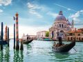 Una scena iconica di Venezia. In primo piano ci sono due gondole con gondolieri che remano attraverso un canale. Sullo sfondo si vede la Basilica di Santa Maria della Salute, un famoso edificio barocco con una grande cupola. L'acqua del canale è verde e riflette il cielo azzurro con alcune nuvole. Ci sono anche pali di ormeggio a strisce arancioni e nere che emergono dall'acqua