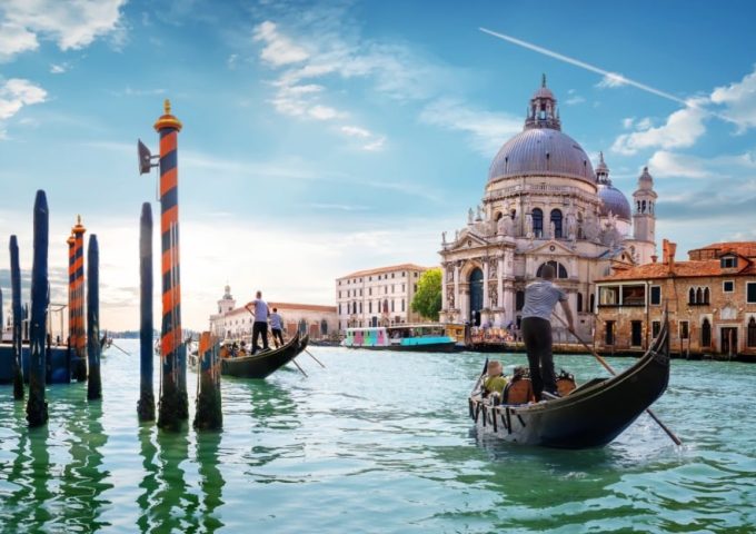 Una scena iconica di Venezia. In primo piano ci sono due gondole con gondolieri che remano attraverso un canale. Sullo sfondo si vede la Basilica di Santa Maria della Salute, un famoso edificio barocco con una grande cupola. L'acqua del canale è verde e riflette il cielo azzurro con alcune nuvole. Ci sono anche pali di ormeggio a strisce arancioni e nere che emergono dall'acqua